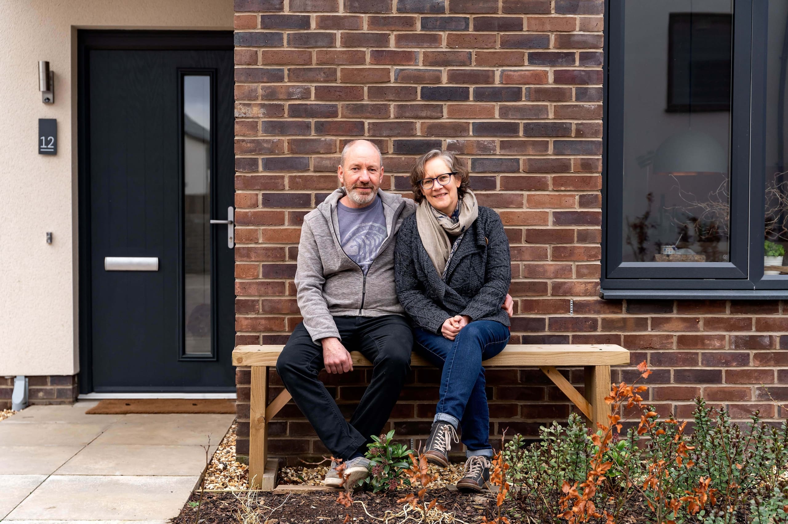 Couple sitting on wooden bench outside a new build house in the Scottish Borders. Dark grey front door in background and brick facade of house behind the couple