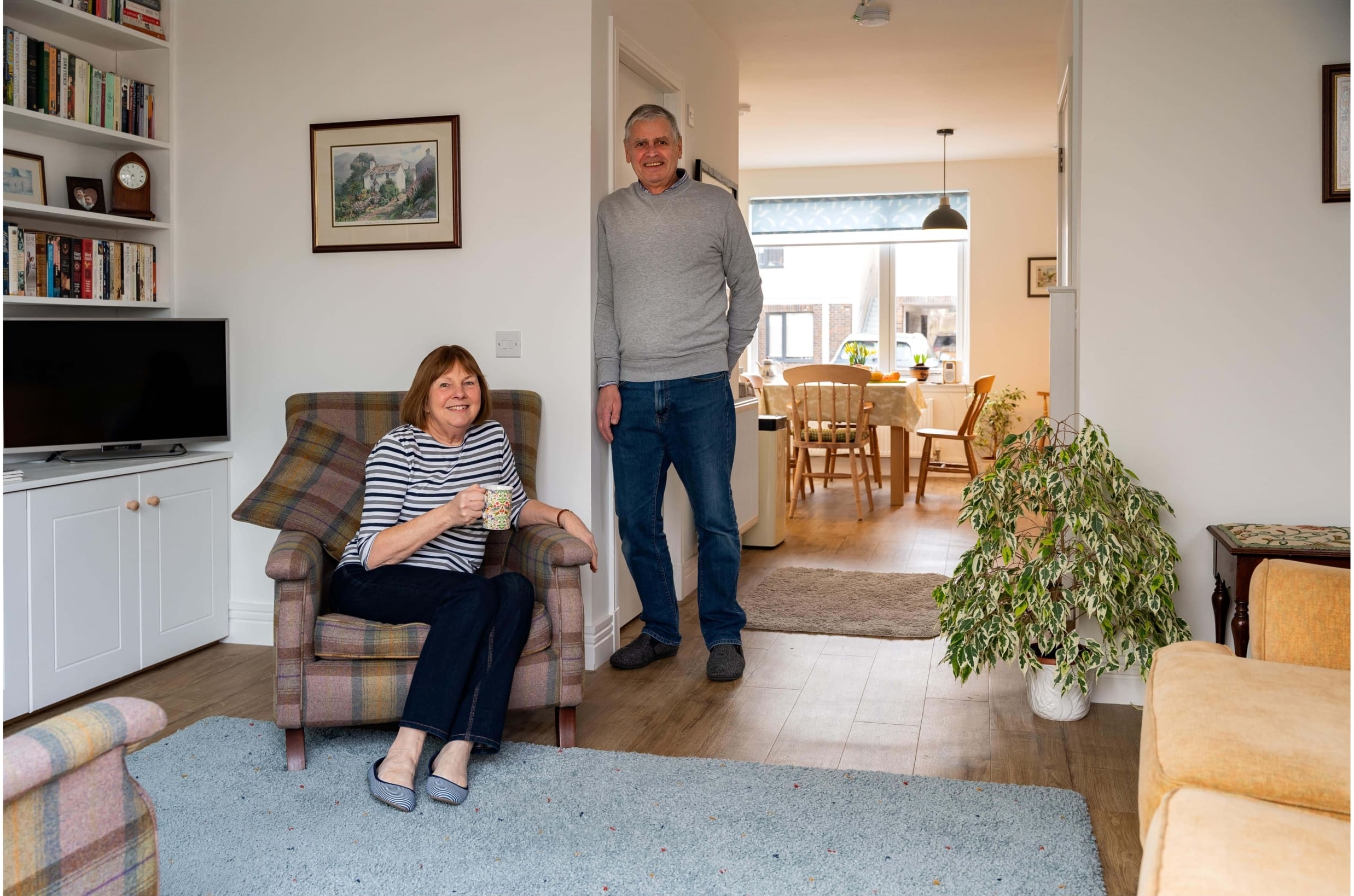 Couple in livingroom that opens into open-plan kitchen/dining area of a new build house in the Scottish Borders. Lady sat on armchair with a mug in hand and man standing, leaning against wall