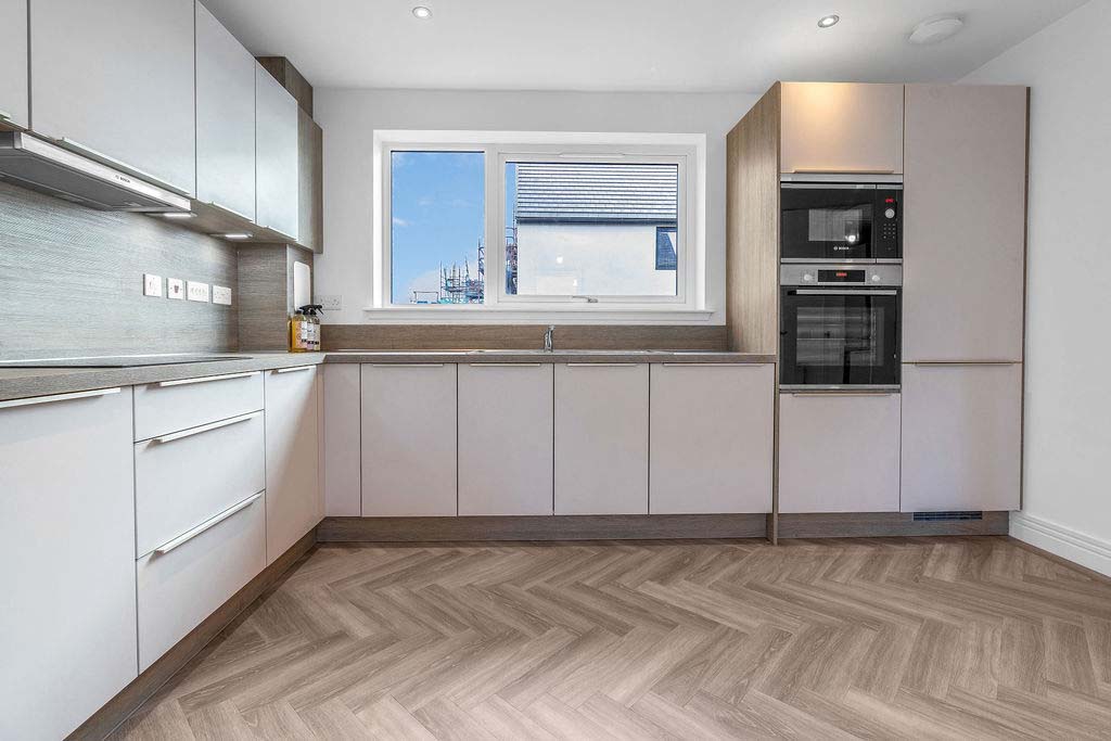 Kitchen along left wall and wall ahead with a large window. Light coloured cupboards and integrated appliances with herringbone wood-effect flooring in a new house for sale in Peebles