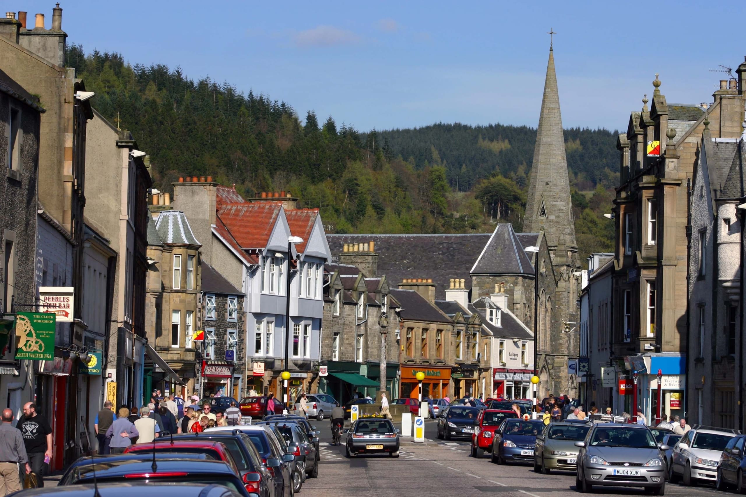 View down highstreet in Peebles. Cars parked to roadside and sandstone buildings with a variety of independent shops