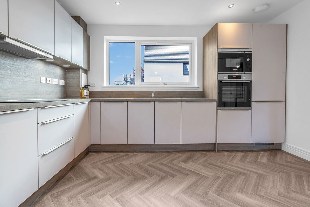 Kitchen along left wall and wall ahead with a large window. Light coloured cupboards and integrated appliances with herringbone wood-effect flooring in a new house for sale in Lauder