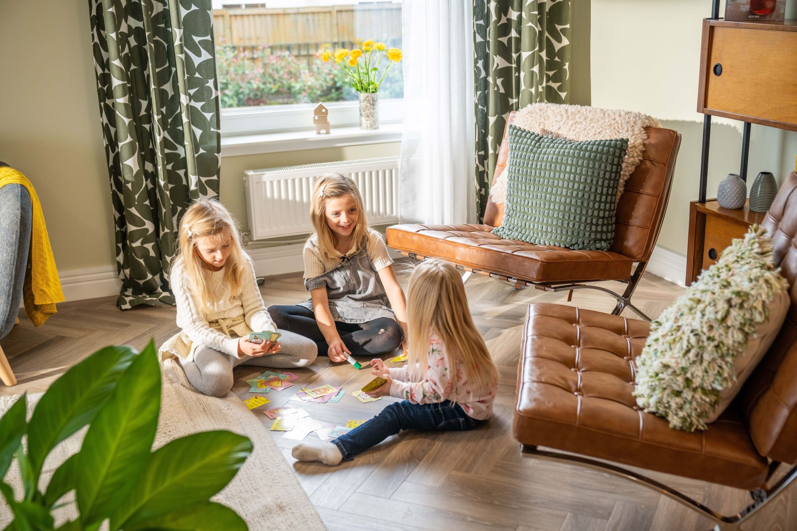 Three girls playing whilst sitting o the floor near a window and two brown chairs with cushions on in a new home for sale in Lauder